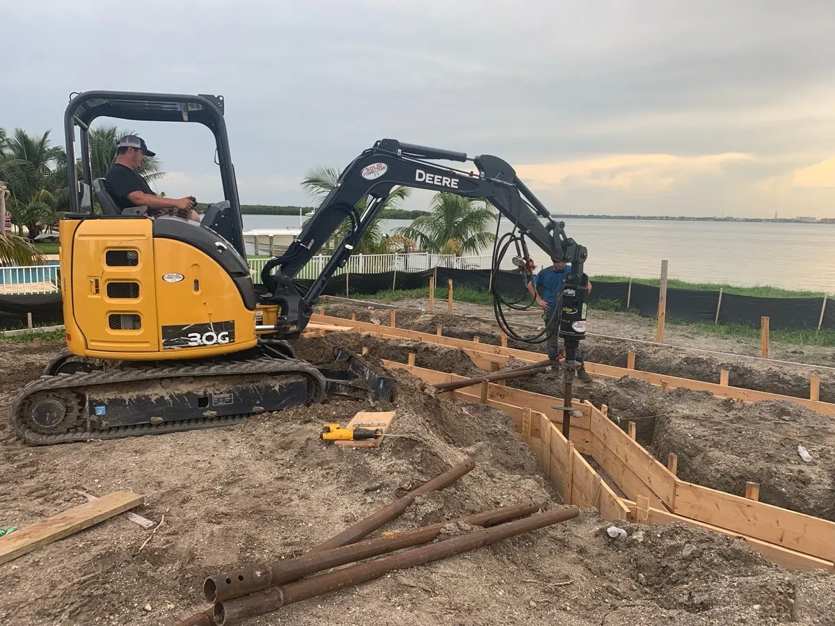 Mini excavator installing helical piers for a waterfront foundation